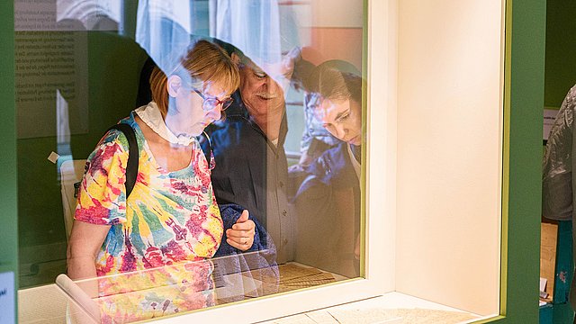 Durch eine rechteckige Vitrine mit grünem Rahmen hindurch fotografierte Besucherinnen, die sich an die Exponate heranlehnen