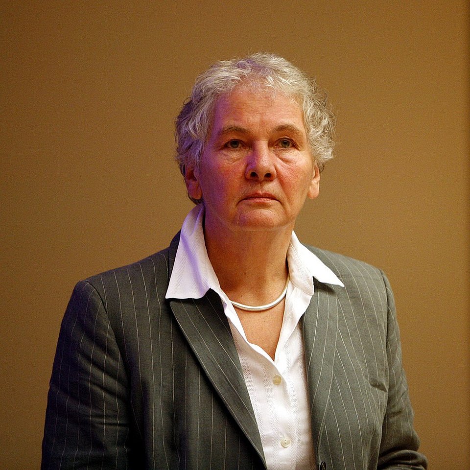 Porträt einer älteren Frau mit kurzen, grauen, lockigen Haaren. Sie trägt einen grauen Nadelstreifen-Blazer über einer weißen Bluse und eine silberne Halskette. Der Hintergrund ist einfarbig in einem warmen Braunton gehalten. Die Frau schaut ernst und nachdenklich geradeaus.