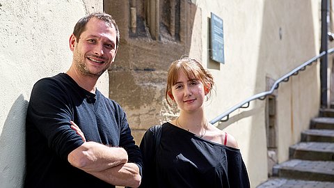 Michael La Corte (links) und Frida Dähn (rechts) stehen vor der sonnenbeschienenen Fassade des Pfleghofs in Tübingen, rechts im Bild führt die Treppe hinauf in Richtung Eingang