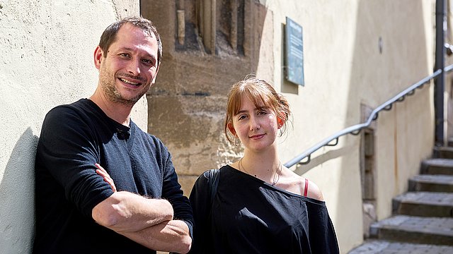 Michael La Corte (links) und Frida Dähn (rechts) stehen vor der sonnenbeschienenen Fassade des Pfleghofs in Tübingen, rechts im Bild führt die Treppe hinauf in Richtung Eingang