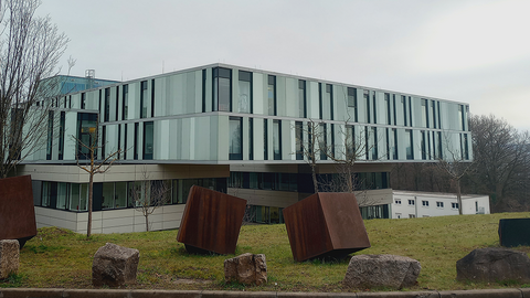 In Hanglage senkt sich vor dem Betrachtenden ein rechteckiges Gebäude mit glaslastiger Fassade hinter einem grünen Rasenstück mit rostbraunen Würfeln