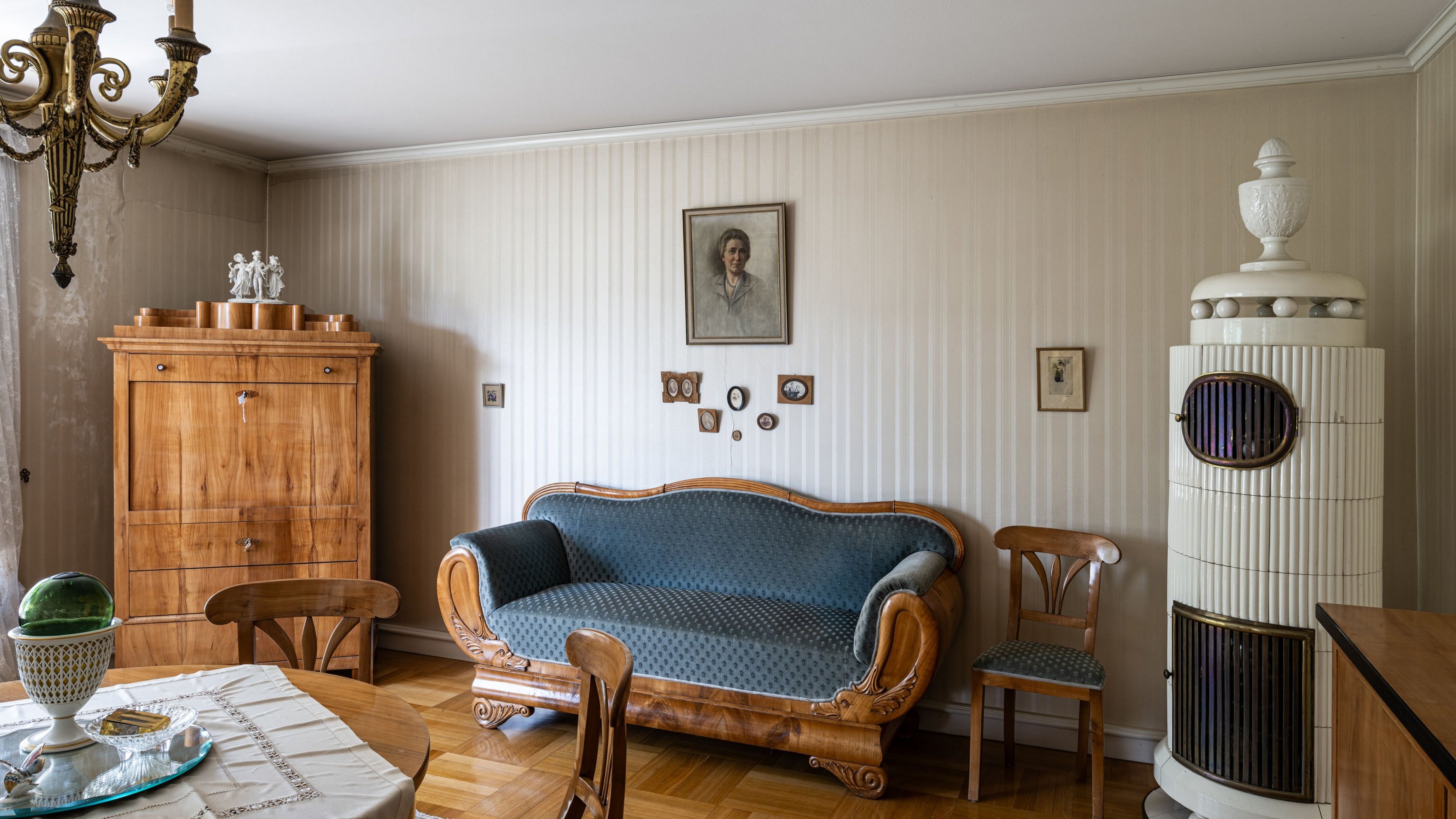 Blick in ein biedermeierlich eingerichtetes Zimmer mit gestreifter Tapete, einem Kachelofen, einem blau bezogenem, geschwungenen Sofa und einem eingeklappten Sekretär.