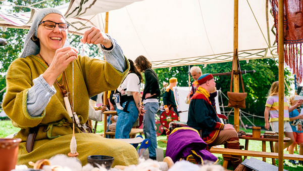 Dame im gelben Kleid, mit weißem Schleier und einer Brille bedient mit den Händen eine herabhängende Spindel, um Garn herzustellen