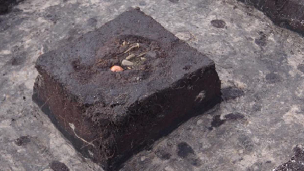 Foto: Prof. Dr. Franz Schopper Rechteckiger, schwarzer Block aus erdähnlichem Material mit mittiger Vertiefung, in der Einschlüsse wie Metallnägel zu sehen sind, in einem dunklen, grauen Grabungsschnitt