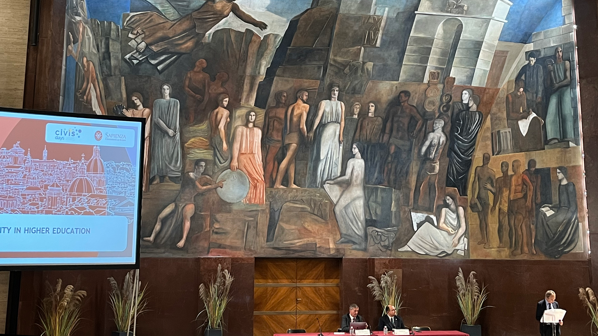 Foto: MUT | Annika Vosseler Begrüßungsveranstaltung der CIVIS Days 2025 in Rom; Hinter dem rot gedeckten Rednerpult ist ein imposantes Gemälde mit einem über einer Menschenmenge fliegenden Engel. Von links ragt ein Bildschirm mit der Titelfolie der CIVIS Days hinein