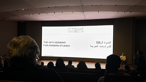 Verdunkelter Raum, beleuchtet von einem großen, zentralen Bildschirm mit dem Titel "The 58th Seminar for Arabian Studies" in Englisch und Arabisch, im Vordergrund die Köpfe des Publikums als schwarze Silhouetten
