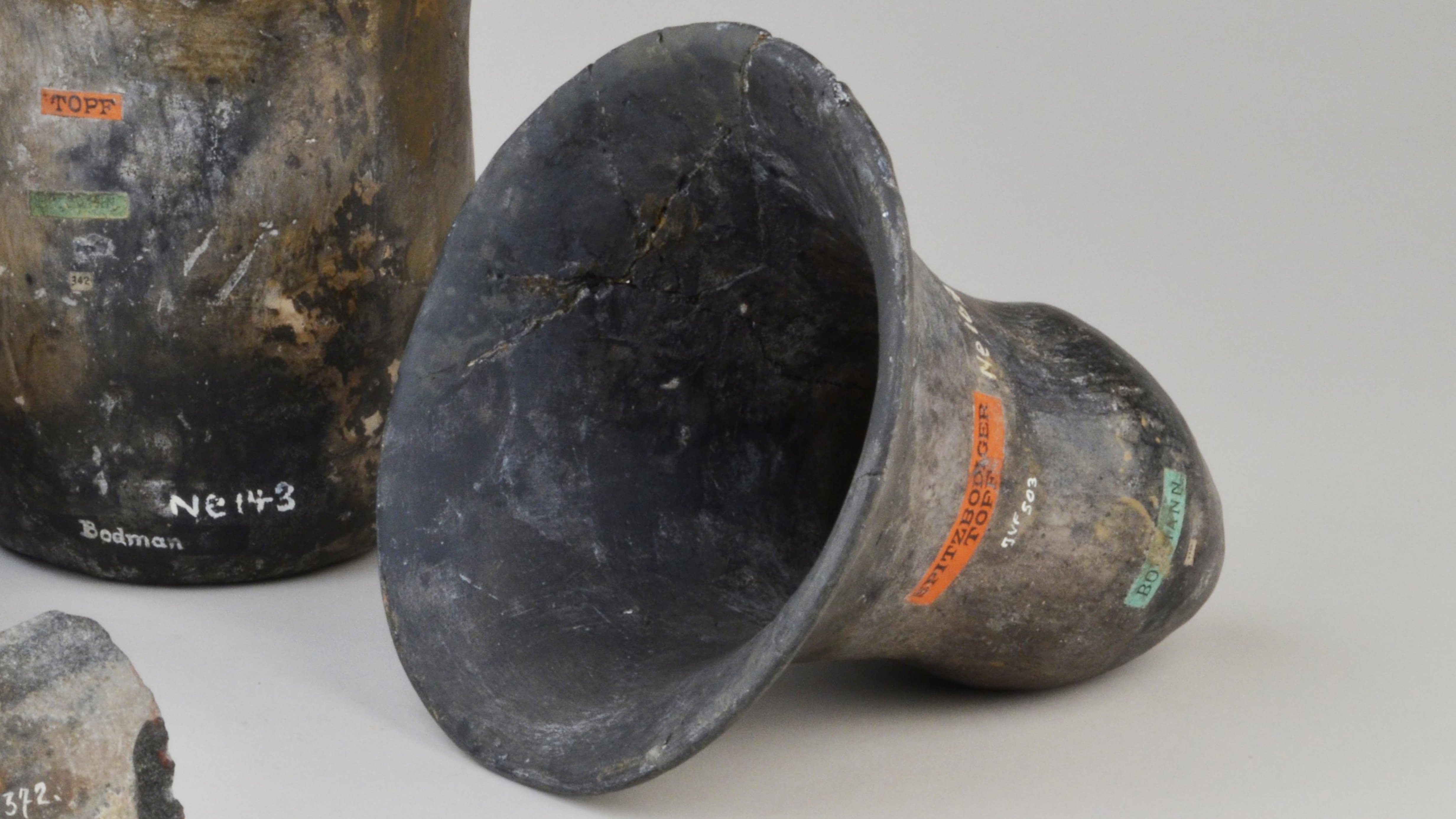 Three pieces of dark ceramic, from left to right: a bulbous jug, a slightly uneven mug, and a bell-shaped vessel lying on its side; in front of them lies an elongated stone piece