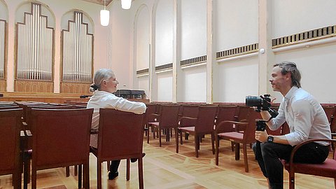 In der mit vielen Sitzreihen bestuhlten Alten Aula der Universität Tübingen sitzt Frau Rektorin Prof. Dr. Karla Pollmann mit dem Arm über der Rückenlehne in der letzten Stuhlreihe und spricht in die von Alexander Ries (rechts) gehaltene Kamera. Im Hintergrund ist die Orgel der Alten Aula zu sehen