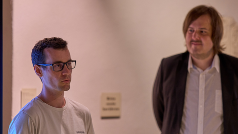 Foto: MUT | Fabian Kurze Links ist im Dreiviertelportrait Luca Dreiling mit Brille und kurzen, dunklen Haaren in einem weißen T-Shirt zu sehen, rechts im Hintergrund unscharf Kevin Körner in weißem Hemd und schwarzer Anzugjacke