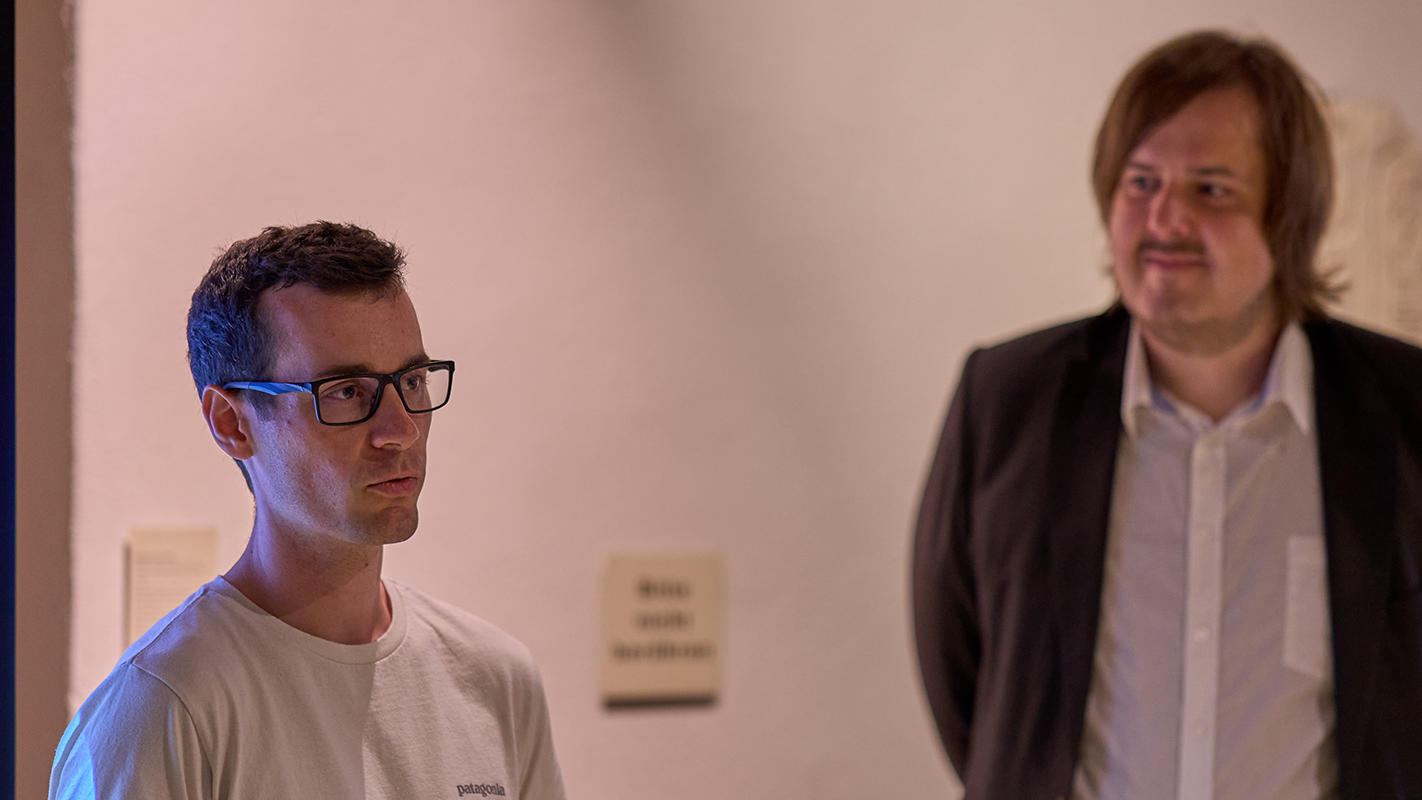 Links ist im Dreiviertelportrait Luca Dreiling mit Brille und kurzen, dunklen Haaren in einem weißen T-Shirt zu sehen, rechts im Hintergrund unscharf Kevin Körner in weißem Hemd und schwarzer Anzugjacke