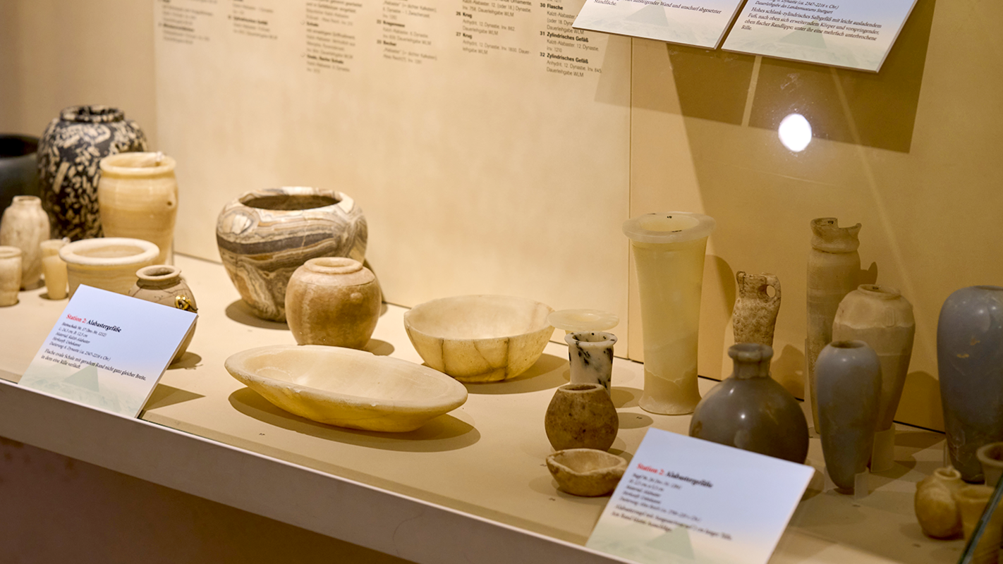 Schräger Blick auf eine Standvitrine mit verschiedenen Formen von Kleinkeramik in Erdtönen, von flachen, ovalen Schalen bis hin zu hohen, schmalen Gefäßen.