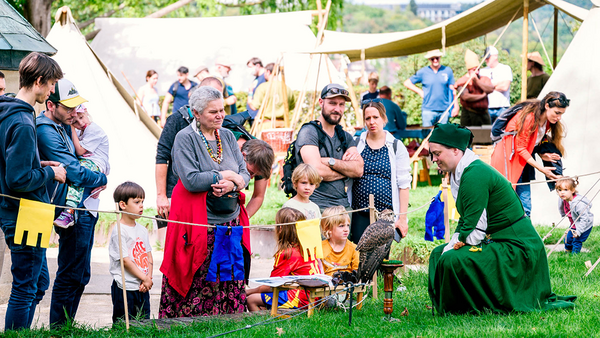 Rechts im Bild eine Dame im grünen Kleid, die neben einer Sitzstange mit einem lebenden, kleinen Raubvogel hockt, auf der anderen Seite eines kleinen Seilzauns sitzen Kinder mit ihren stehenden Begleitpersonen