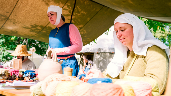 Niedriger, schräger Blick auf einen Tisch mit gefärbten Wollbeispielen, hinter denen zwei Damen in gelber beziehungsweise blau-rosaner Kleidung mit weißen Hauben sitzen