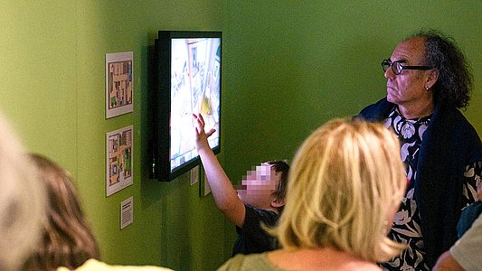 Ein Touchscreen an grün gestrichener Wand, zu dem ein Junge hinaufreicht, im Vordergrund sind Besuchende zum Teil zu erkennen