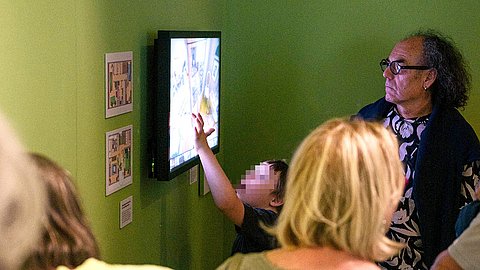 Ein Touchscreen an grün gestrichener Wand, zu dem ein Junge hinaufreicht, im Vordergrund sind Besuchende zum Teil zu erkennen