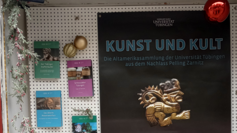 Foto: MUT | Nadja Mozdzen Rückwand der Vitrine, auf der links die kleinen Monographien und rechts das Poster zur Ausstellung "Kunst und Kult" mit einem goldenden, figürlichen Ornament angebracht sind; links hängt eine Kette aus falschem Immergrün