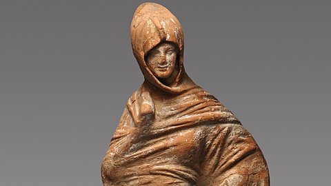 Foto: MUT | Valentin Marquardt Terrakotta-Statuette einer in ihren Mantel gehüllten Frau, die eine Hand in die Hüfte stemmt und die andere in ihrem Mantel verbirgt