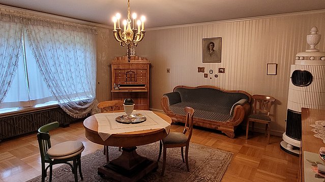 Wohnzimmer im Biedermeier-Stil mit Blick auf ein Sitzensembleunter einem Kronleuchter, zur Linken Gardinen vor dem Fenster und an der Wand Sofa, Keramikofen und Schrank
