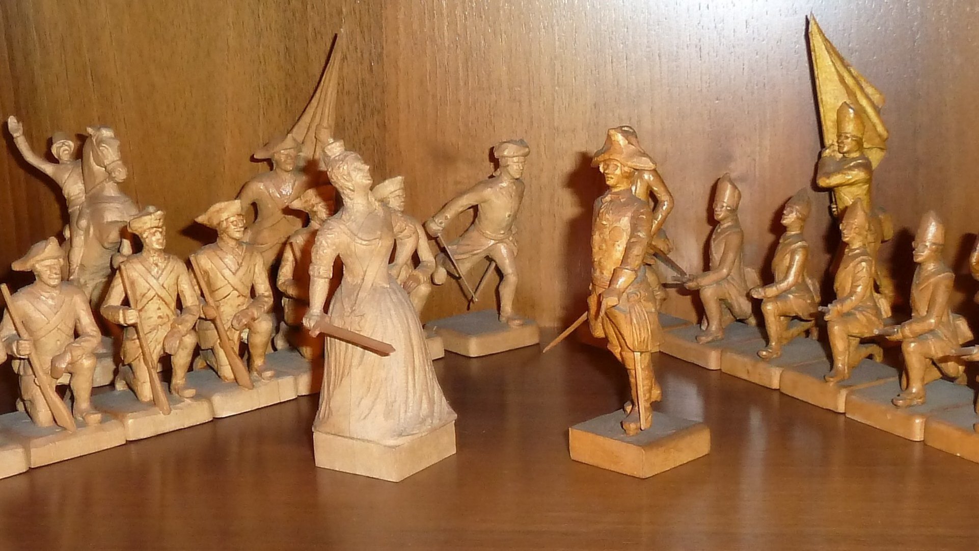 Im Winkel aufgestellte, aus Holz geschnitzte Schachfiguren: links die hellen Bauern mit einem Springer, rechts die dunklen Bauern mit einer fahnendentragenden Figur; in der Mitte stehen die helle Königin und der dunkle König einander gegenüber.
