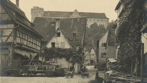 Historische Photographie einer Kreuzung zweier schmaler, gepflasteter Straßen mit Fachwerkhäusern mit steilen Dächern darum herum, im Hintergrund erhebt sich Schloss Hohentübingen; darunter steht handschriftlich notiert "A. von der Trappen, 3959, Tübingen, Seelhausgasse am Schloss"