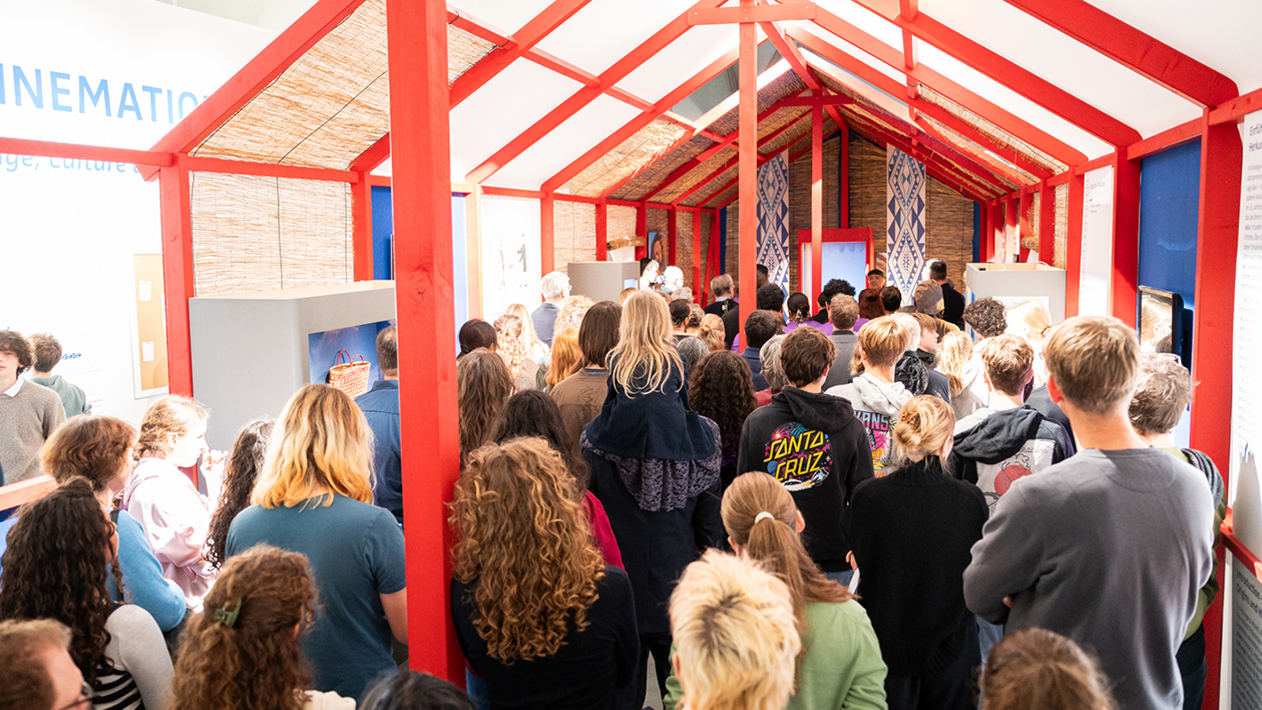 Rückwärtige Ansicht der Menge an Besuchenden, die sich während der Zeremonie im stilisierten Versammlungshaus des Ausstellungsaufbaus versammelt haben