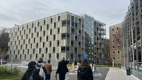 Prof. Dr. Seidl mit einer Gruppe Studierender am unteren Bildrand in der Rückansicht; er zeigt auf das Parkhaus der Kliniken Tal, einen achtstöckigen Bau Stahlbau mit einem eingesetzten gläsernen Treppenhaus