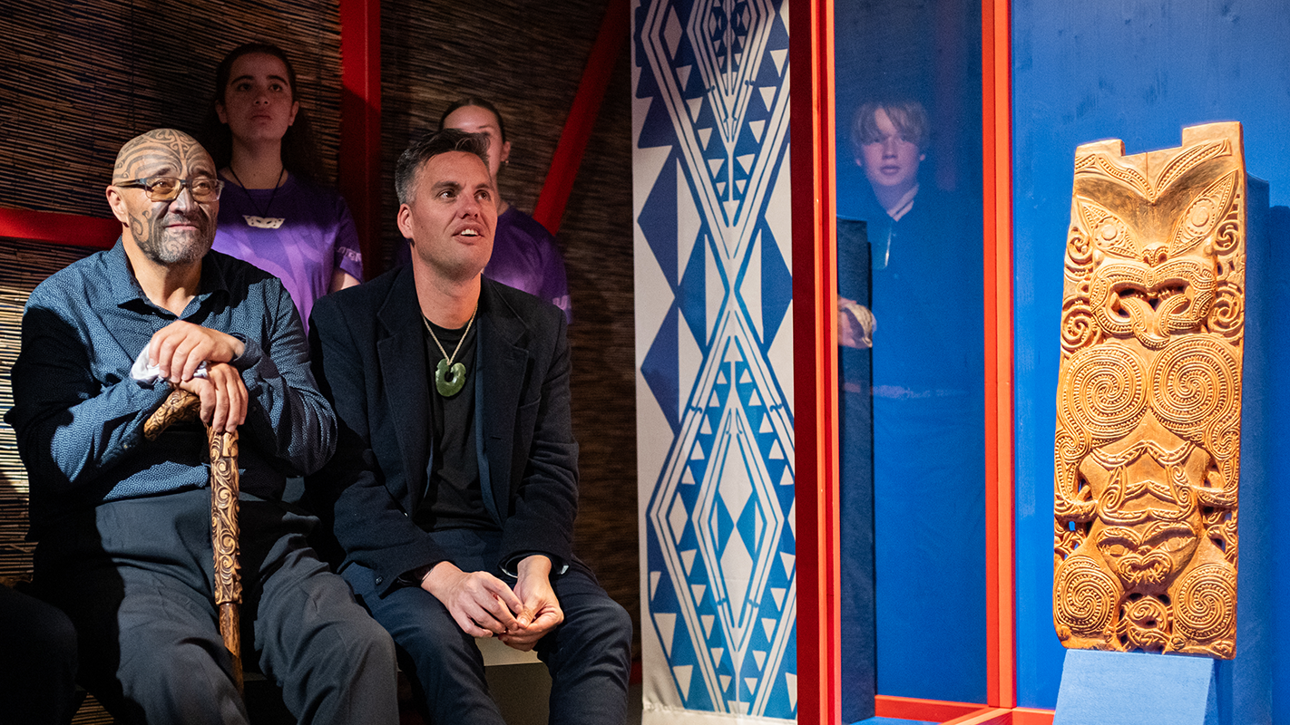 Zur Linken sitzen zwei Teilnehmende der Zeremonie aus verschiedenen Maori-Communities, zur Rechten ist die erleuchtete Vitrine mit dem Pou von Hinematioro