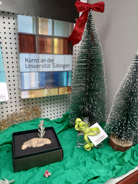 Foto: MUT | Nadja Mozdzen Hochkant eine Vitrinenecke mit zwei kleinen Tannenbäumchen aus Draht rechts im Bild, zur Linken liegt ein schwarzes Schächtelchen mit einer elfenbeinfarbenen Löwenreplik und ein Ausstecherle in Form des Urpferdchens; im Hintergrund ist auf einem hängenden Ständer das Buch "Kunst an der Universität Tübingen" zu sehen