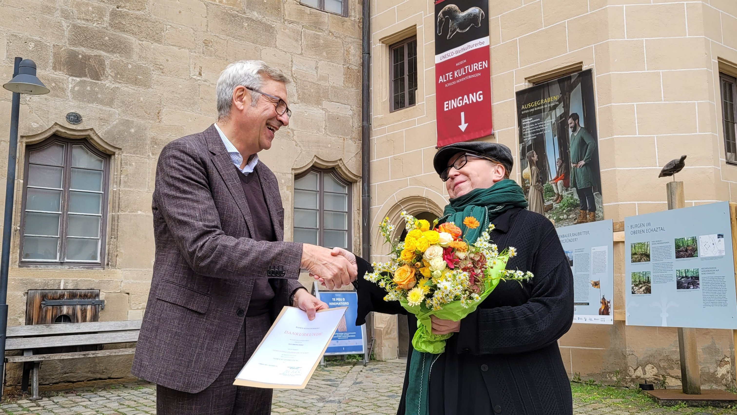 Museumsdirektor Prof. Dr. Ernst Seidl (links) und Christina Häfele M.A. (rechts) stehen vor dem Eingang des Museums Alte Kulturen auf Schloss Hohentübingen; er überreicht ihr eine Urkunde und schüttelt ihr die Hand, sie hält einen bunten Blumenstrauß