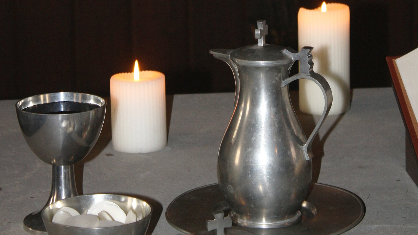 Liturgical equipment made from silver; a chalice, pitcher, and bowl with white wafers; two burning candles in the background