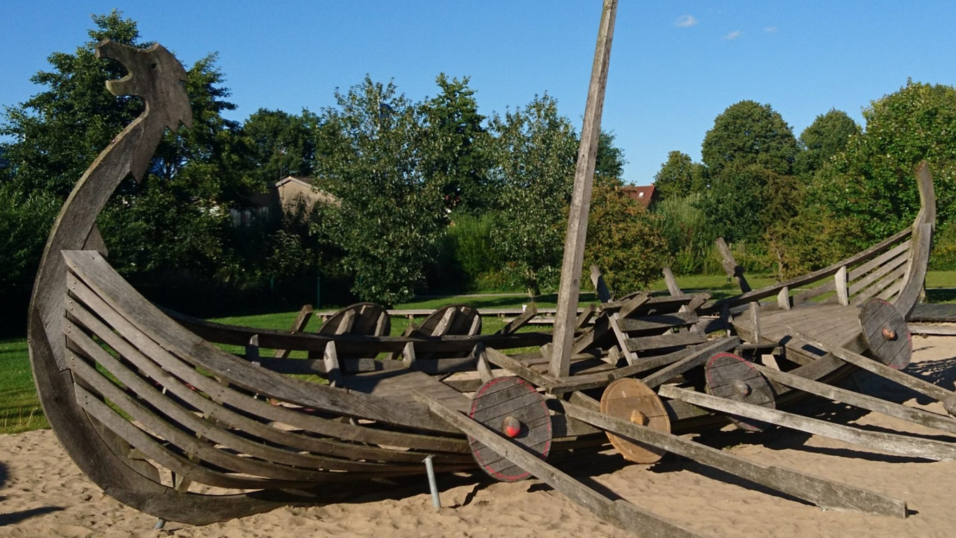 Foto: Universität Tübingen Ein hölzernes Klettergerüst in Form eines Landschiffes in einem Sandkasten, im Hintergrund grüne Bäume und blauer Himmel.