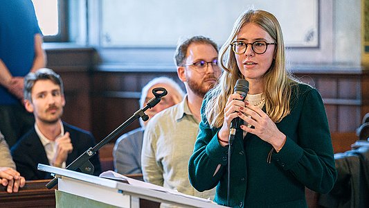 Teresa Pohl im petrolfarbenem Oberteil hält frei das Mikrofon und spricht nach schräg links gewandt, hinter ihr steht zuhörend Lucas Rau