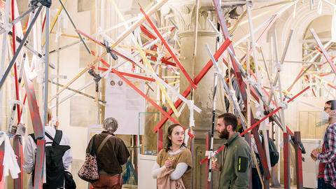 Besuchende stehen einzeln oder in Gruppen in der Ausstellung in den drei Erkern im Rittersaal, aus denen das Konstrukt aus verschiedenfarbigen Holzstangen herausragt