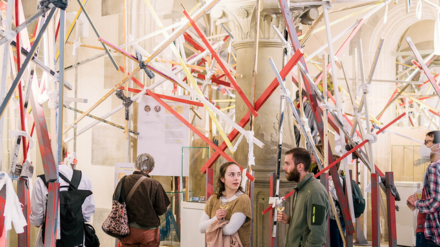 Besuchende stehen einzeln oder in Gruppen in der Ausstellung in den drei Erkern im Rittersaal, aus denen das Konstrukt aus verschiedenfarbigen Holzstangen herausragt
