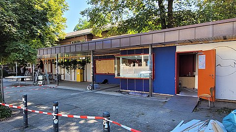Außenansicht des sommerlichen Unipavillons mit Absperrband im Vordergrund, Holzmaterial rechts im Bild und weit geöffneten Türen