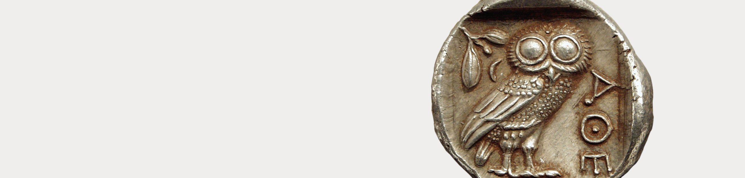 Nahaufnahme einer silbernen Tetradrachme mit seitlich stehender Eule, Olivenzweig und "ATHE" auf hellgrauem Grund