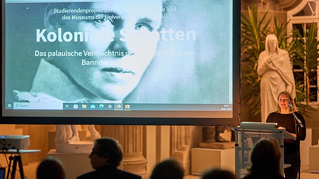 Dr des Annika Vosseler am Rednerpult der Eröffnung „Koloniale Schatten“; auf der Leinwand links im Bild das Titelbild mit dem Schwarz-Weiß-Portrait von Elisabeth Krämer-Bannow und dem Schriftzug des Titels
