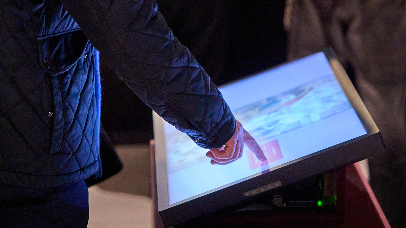Schräg auf einem Tisch montierter Touchscreen mit einer interaktiven Überlagerung eines Medici-Gemäldes; ein Besuchender berührt mit dem Zeigefinger die Anzeige