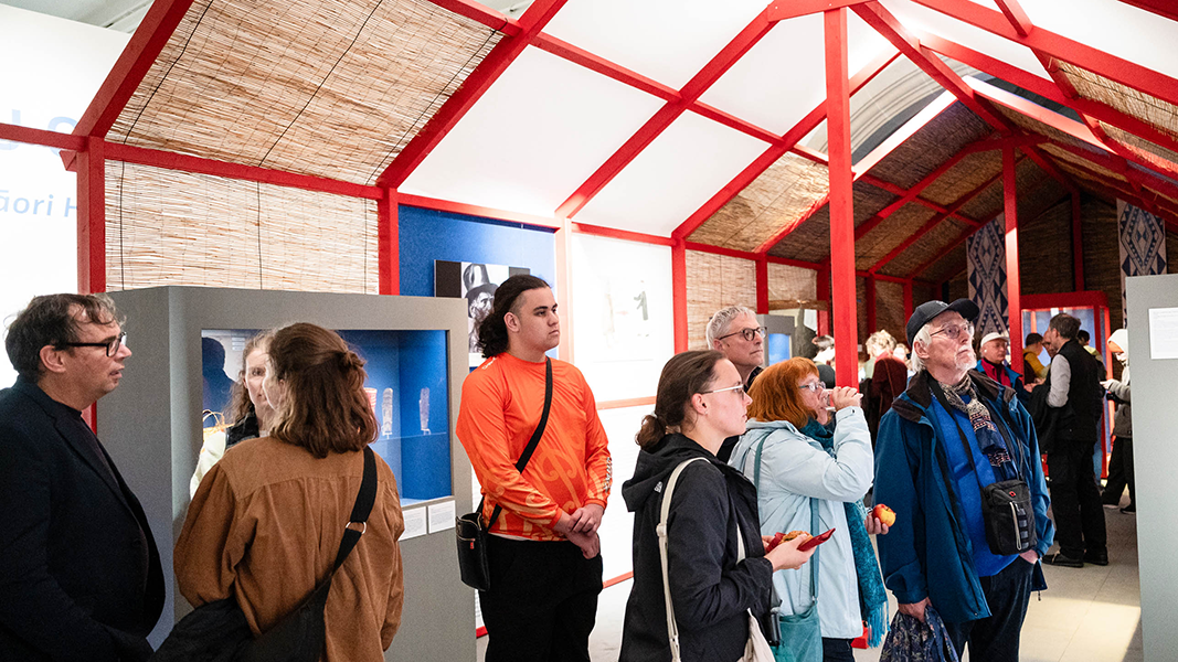 Eine Reihe Besuchender, unter ihnen ein Schüler im orangenen Shirt der Delegation der Te Aitanga-a-Hauiti, steht im abstrakt errichteten Langhaus und betrachtet eine Wand der Ausstellung, die nicht im Bild zu sehen ist