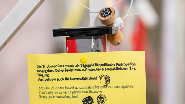 Nahaufnahme eines Stempels, der mit einem Nylonfaden im Gestänge angebunden ist und auf einem geschlossenen Stempelkissen ruht, das Motiv zeigt eine Münzprägung mit Eule, darunter ist ein gelbes Schild mit sauberer Handschrift, das den Stempel als Triobol-Münz-Symbol benennt, nach der Silbermünze in Höhe des Tagegeldes der athenischen Demokratie im 4. Jahrhundert v. Chr.