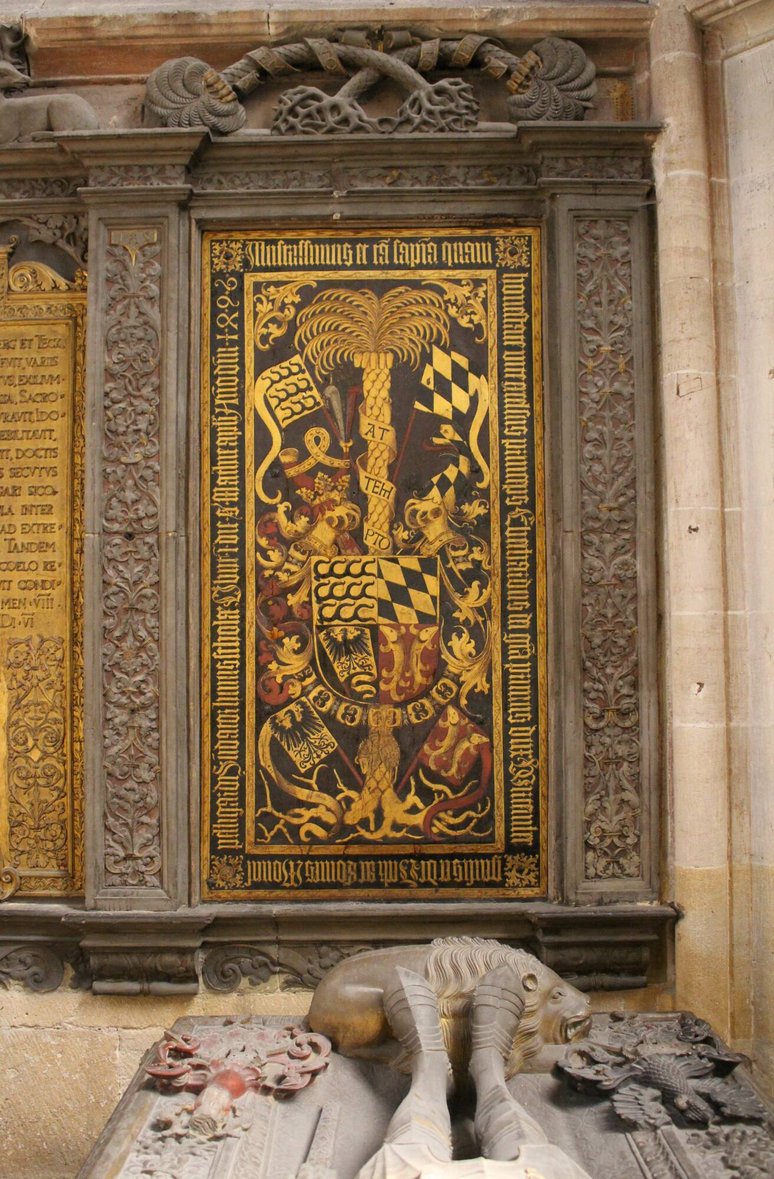 Innenaufnahme eines prunkvoll verzierten Raumes. Im Mittelpunkt steht eine große, reich mit Gold verzierte Holztür oder Wandverkleidung mit symmetrischen floralen und ornamentalen Mustern. Umgeben ist sie von hellen Wänden mit Stuckelementen. Im unteren Bildbereich sind weiße Stühle mit hohen Lehnen zu sehen, die auf eine museale oder repräsentative Nutzung des Raums hinweisen.