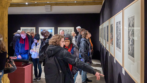 Besuchende in der Ausstellung im Kabinettraum mit schwarz bespannten Wänden; ein Besucher im Vordergrund deutet auf einen der hellen, gerahmten Drucke an der Wand