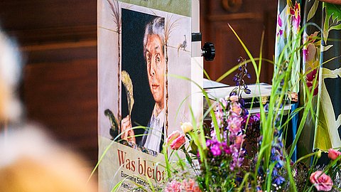 Blick auf einen Ausschnitt des Rednerpults mit frontalem Plakat der Ausstellung, Portrait von Evamarie Sander in der Mitte, mit Blumenstrauß im Vordergrund
