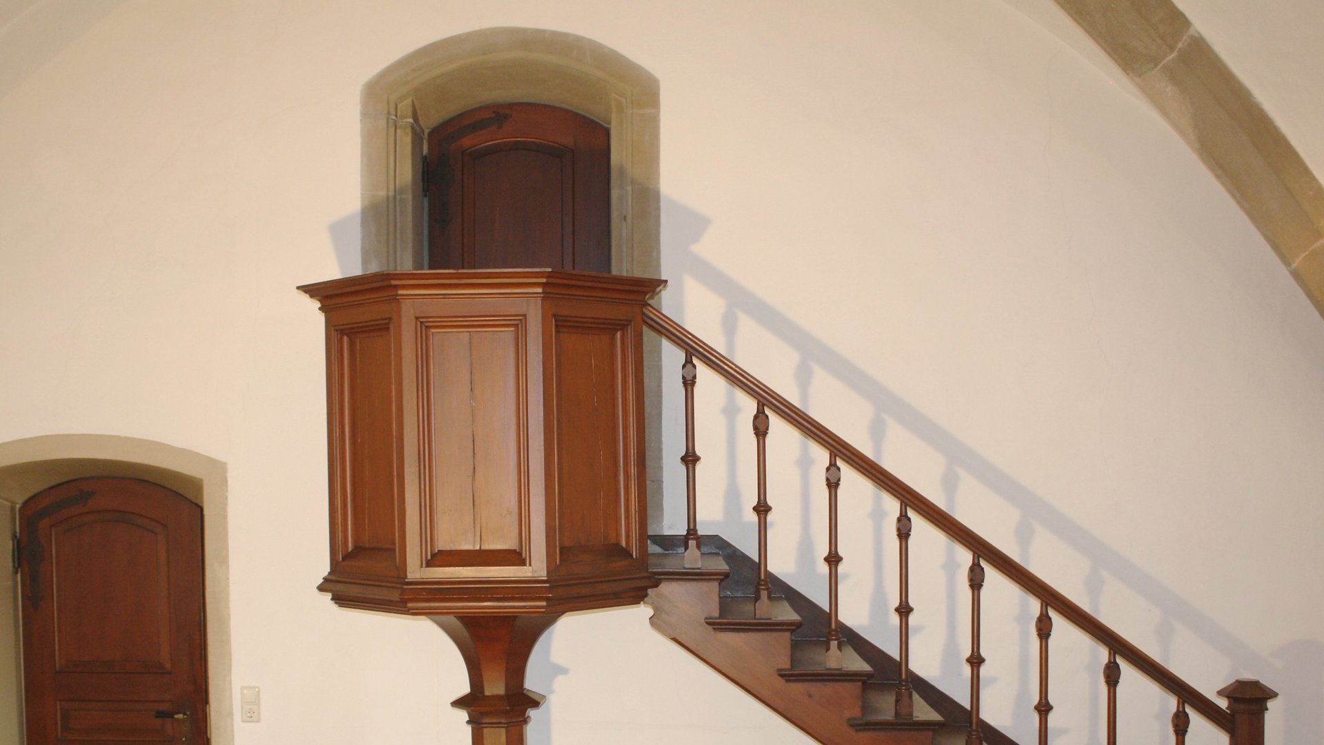 Foto: MUT Weiße Wand mit einer Holztür unten sowie einer erhöhten Holztür mit einem kanzelartig abgeschirmten Absatz davor, zu der eine Treppe hinaufführt
