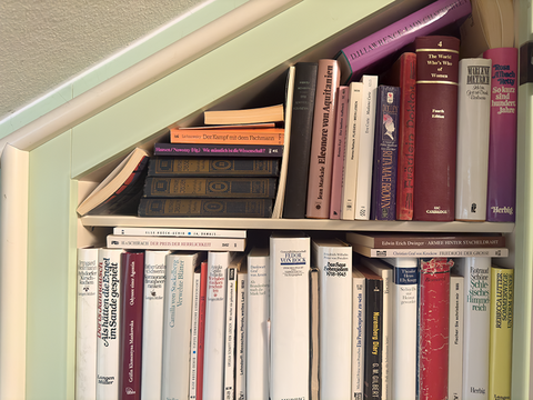Ausschnitt eines Wandschrank-Bücherregals mit der mintgrün gestrichenen Dachschräge links im Bild, gefüllt mit Romanen mit weißen, roten und dunklen bedruckten Buchrücken