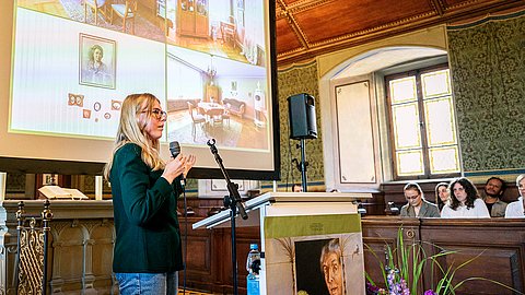 Teresa Pohl spricht, das Mikrofon frei in der Hand, nach rechts gewandt vor dem Rednerpult (rechts) und der Leinwand mit Aufnahmen aus dem Haus Sander (links)