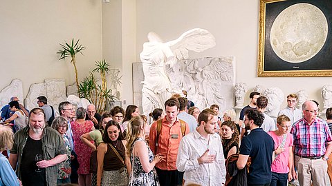 Besuchende der Ausstellungseröffnung stehen in Grüppchen im Rittersaal, mittig hinter ihnen der Abguss der Nike von Samothrake und rechts das Mondgemälde