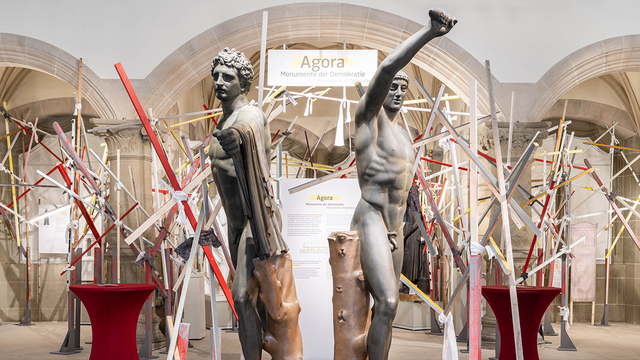 Breite Panorama-Ansicht der Sonderausstellung "Agora" in den Nischen des Rittersaals, mit einem vielfarbigen Gerüst aus Holzbalken unterschiedlicher Größe und im Vordergrund frontal die Tyrannenmörder-Abgüsse mit erhobenen Waffenarmen