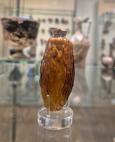 Foto eines gelben Glasfläschchens auf einem transparenten Ständer in einer Glasvitrine der Ausstellung.
