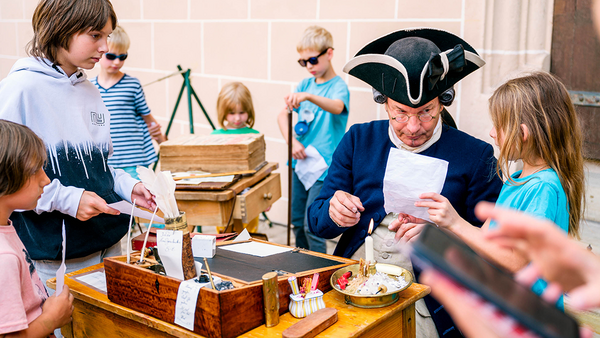Darstellender einer Feldkanzlei mit blauem Uniformrock und Dreispitz, der einem Kind ein handbeschriebenes Stück Papier reicht